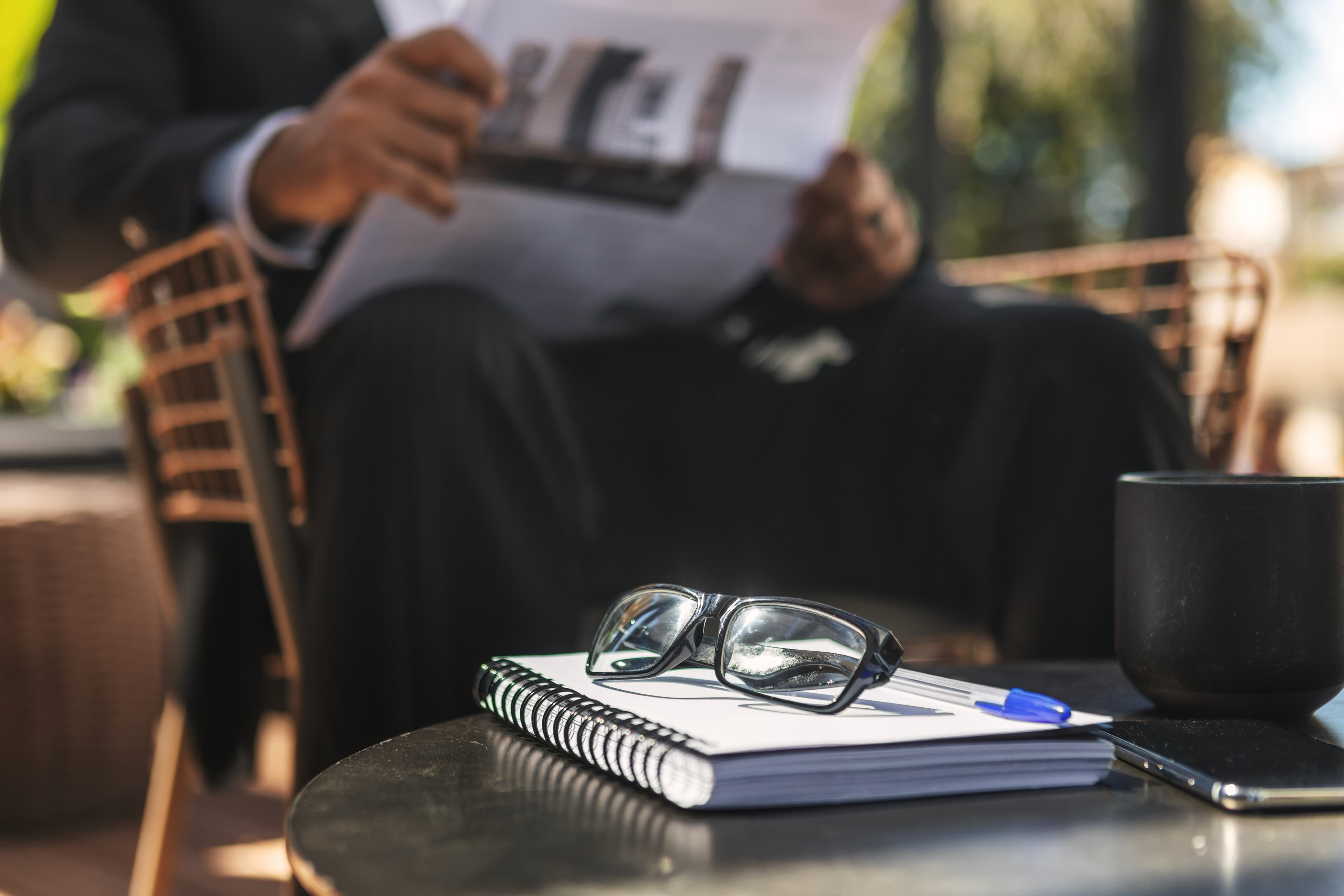 Glasses and a notebook on a table, with someone reading a report in the background