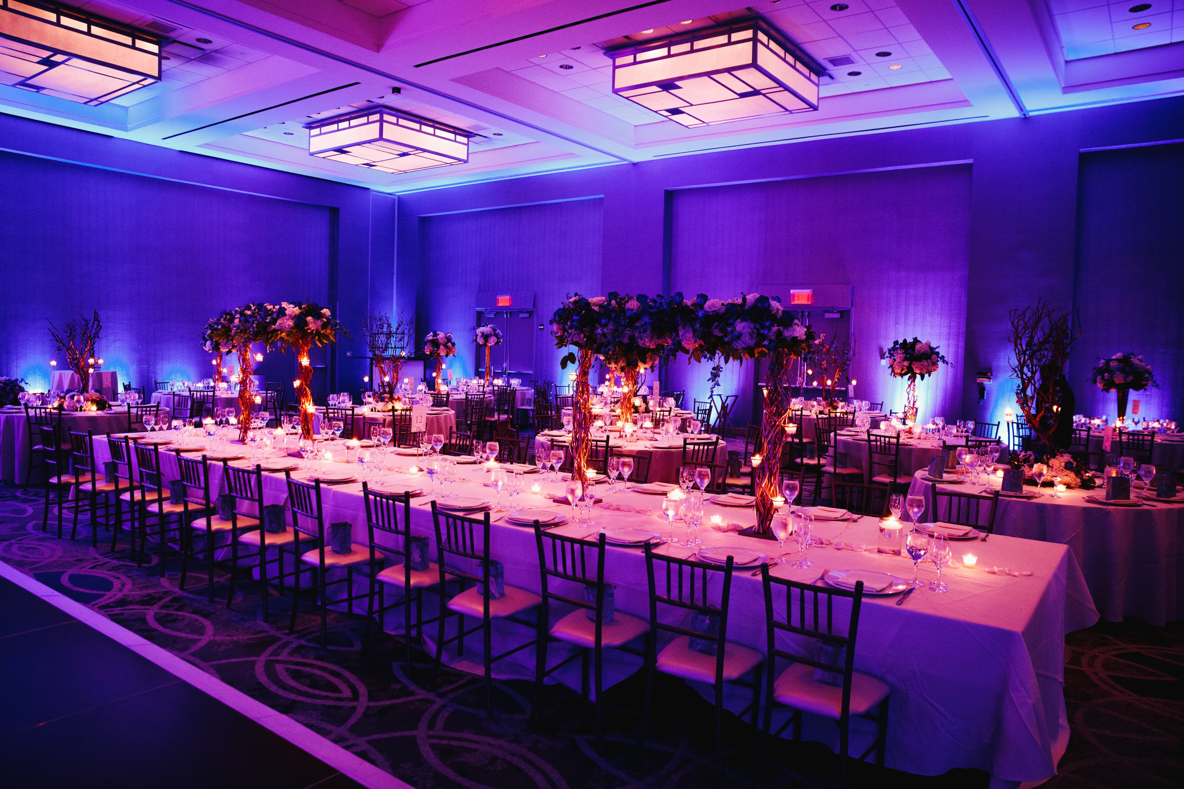 A formally set banquet hall with long tables, floral centrepieces and purple uplighting
